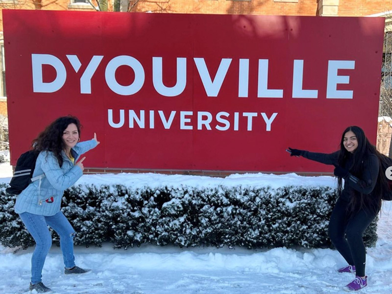 Two female students point to D'Youville campus sign in the snow